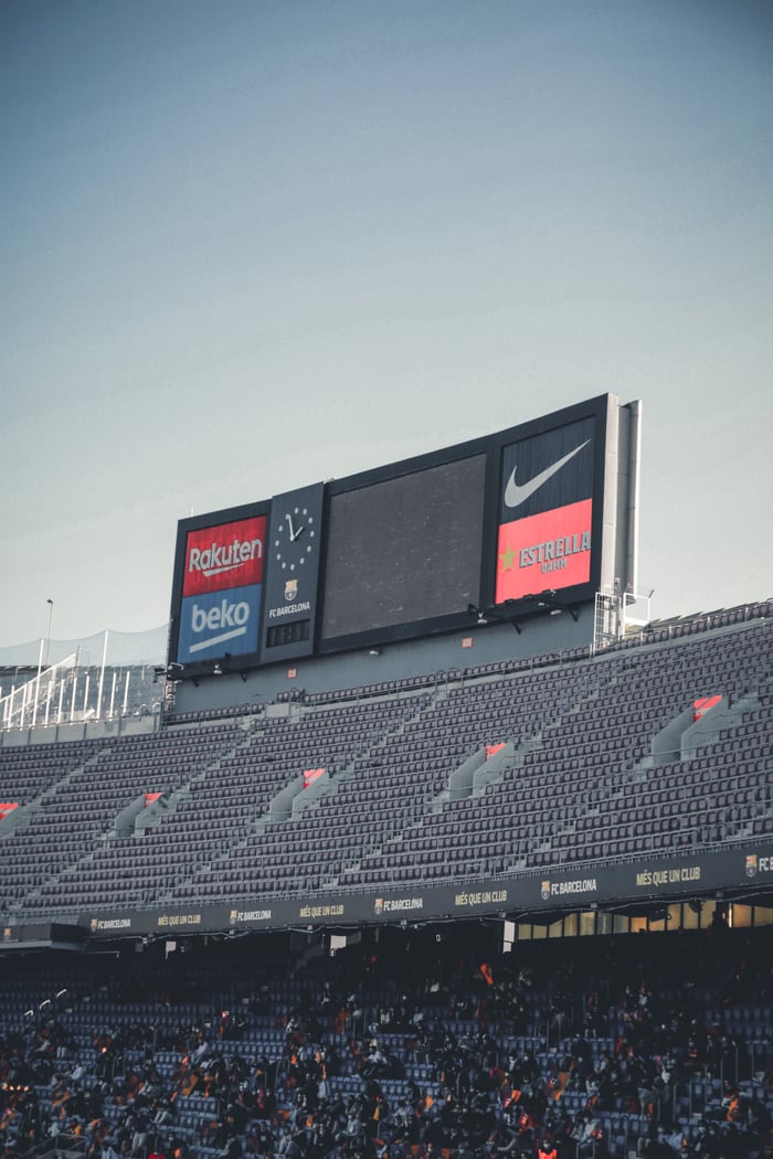 A large scoreboard in a stadium displaying various brand logos under a clear sky.